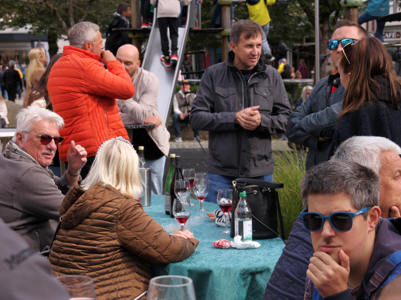 Frauen und Männer sitzen an gedeckten Tischen im Freien und trinken ein Glas Rotwein, andere stehen daneben und sprechen miteinander.