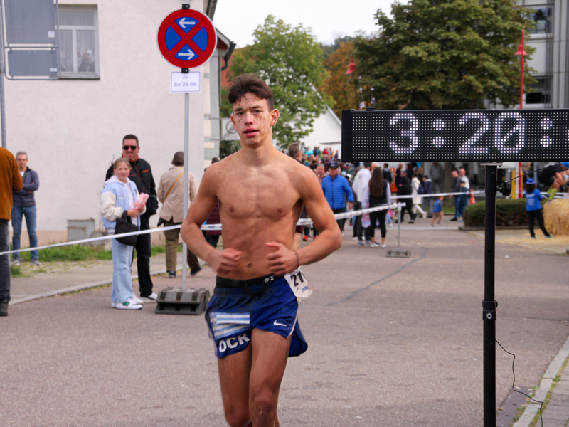 Ein Läufer mit nackigem Oberkörper und Dreckspritzern im Gesicht rennt an einer Zeitmesstafel vorbei, auf der 3:20:4 steht. Passanten stehen an einer Absperrung der Laufstrecke und schauen zu.