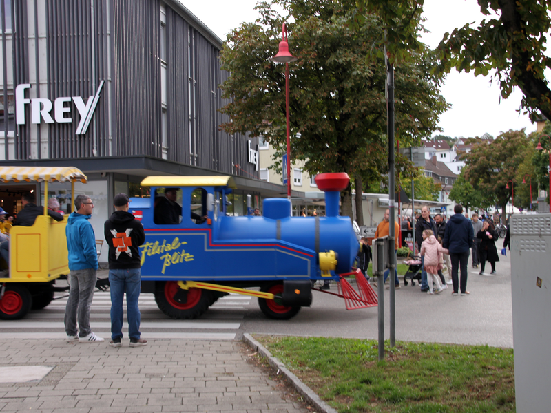 Ein zum Mini-Zug umgebauter Traktor zieht einen Anhänger voller Menschen über eine Straße, vorbei an Passanten und einem Bekleidungsgeschäft.
