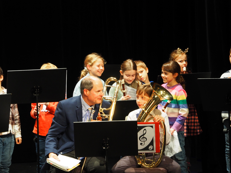 Ein Mann mit einer Posaune sitzt  beim Adventskonzert der Musikschule im Uditorium auf einer Bühne, umgeben von mehrfach lachenden Kindern, die Flöten halten.