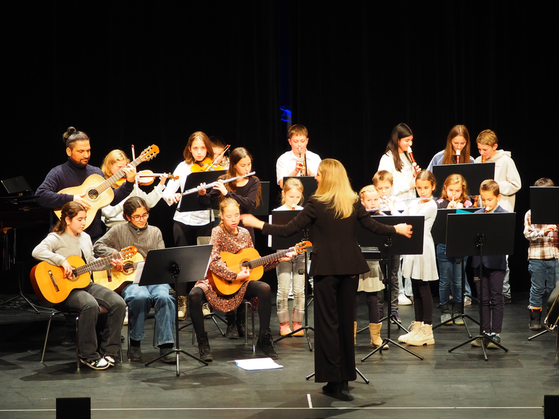 Eine Gruppe von Kindern spielt unter der Leitung einer Dirigentin auf einer Bühne verschiedene Instrumente wie Gitarren, Geigen und Blockflöten beim Adventskonzert der Musikschule im Uditorium.