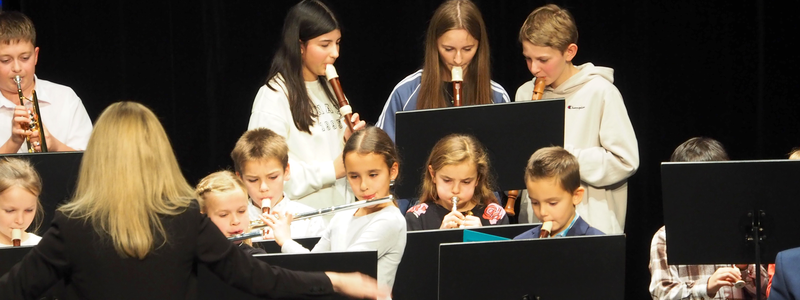 Eine Gruppe von Kindern spielt Blockflöten auf der Bühne des Uditoriums beim Adventskonzert der Musikschule, geleitet von einer Dirigentin.