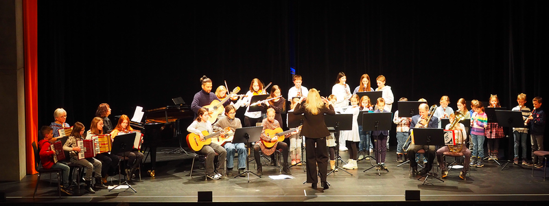 Eine Gruppe von Musikern, darunter Kinder, Jugendliche und Erwachsene, spielt auf der Bühne des Uditoriums vor einem Publikum verschiedene Instrumente beim Adventskonzert der Musikschule. 