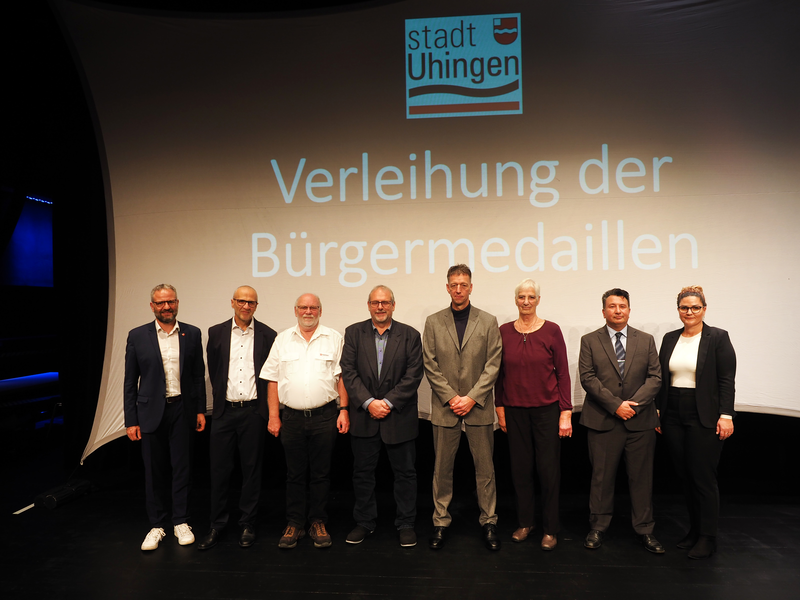 Acht Personen stehen vor einer Leinwand mit der Aufschrift "Verleihung der Bürgermedaillen" und dem Logo der Stadt Uhingen.