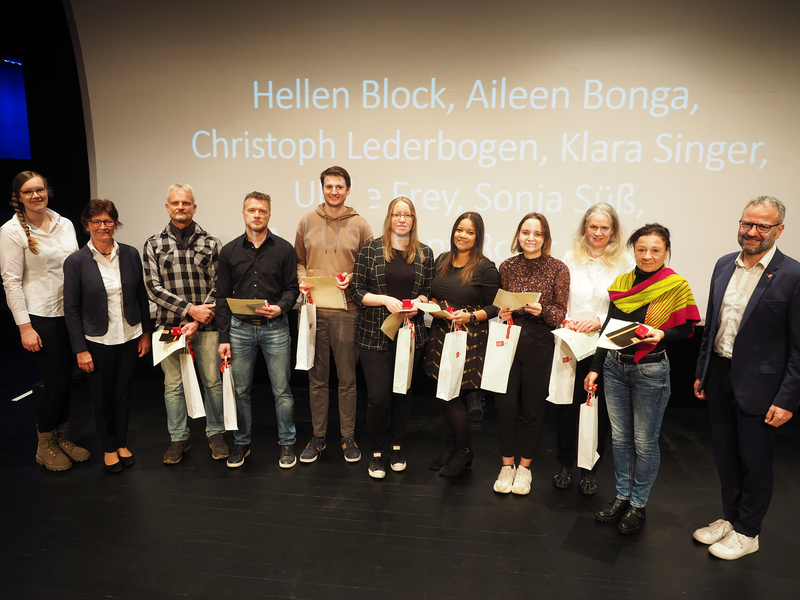 Eine Gruppe von zwölf Personen steht nebeneinander auf einer Bühne und hält Geschenkpakete in der Hand, während hinter ihnen Namen auf eine Leinwand projiziert sind.