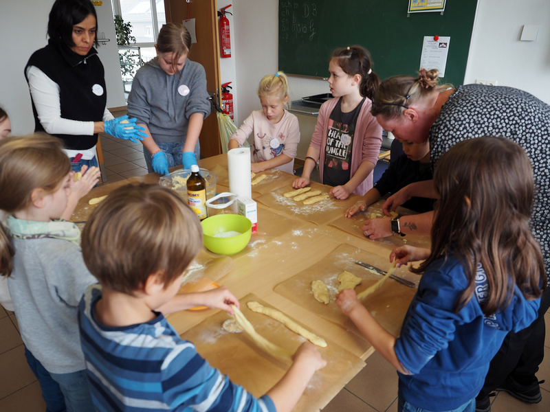 Eine Gruppe von Kindern formt unter Anleitung von Erwachsenen auf einem Tisch Teigstücke für Hefezöpfe.
