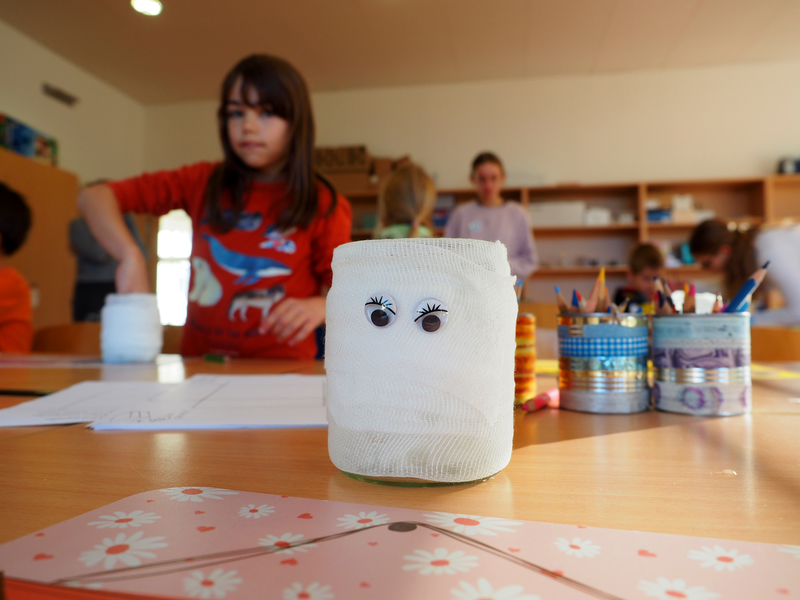 In einem Klassenraum der Grundschule Sparwiesen sitzen Kinder an einem Tisch mit Bastelmaterialien, während ein mit Augen dekoriertes Glas im Vordergrund steht.