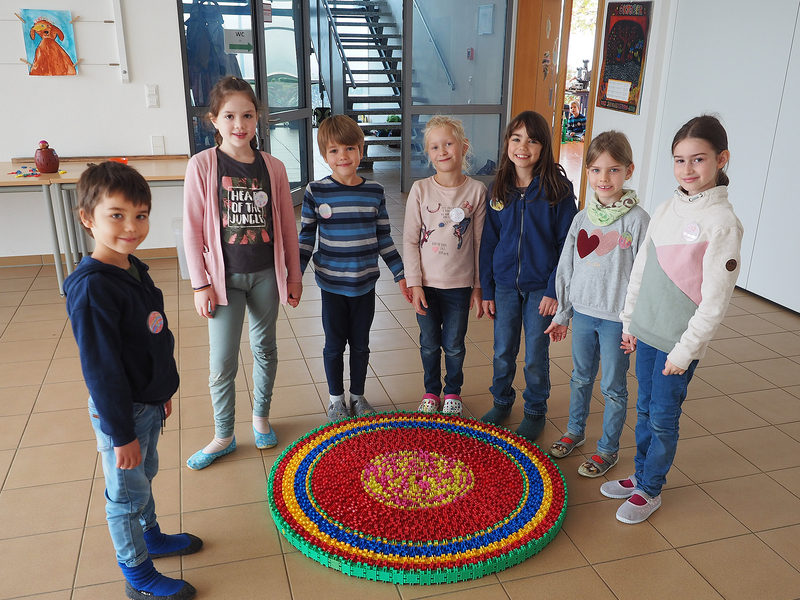 Eine Gruppe von sieben Kindern steht bei der Ferienbetreuung der Stadt Uhingen im Kreis um ein großes, buntes Mandala auf dem Boden im Foyer der Grundschule Sparwiesen.
