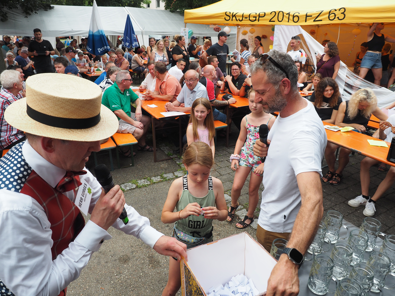 Ein Mädchen zieht aus einer Kiste ein Los, hinter ihr sitzen Männer und Frauen an Biertischen.