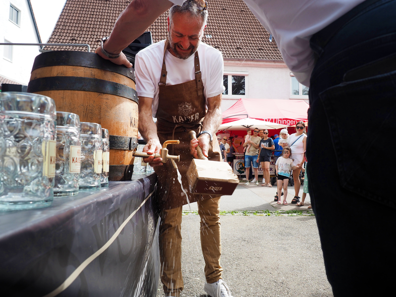 Uhingens Bürgermeister Matthias Wittlinger schlägt mit einem großen, viereckigen Holzhammer einen Zapfhahn in ein Bierfass aus Holz und etwas Bier sprudelt hinaus. Menschen schauen ihm zu.