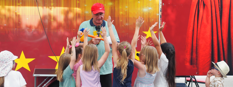 Ein Zauberkünstler steht vor einem bunten Wagen und vor ihm stehen einige Kinder