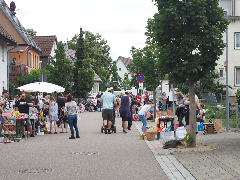 Auf einer Straße stehen bei einem Kinderflohmarkt viele Stände mit Spielsachen und anderen Kinderartikeln
