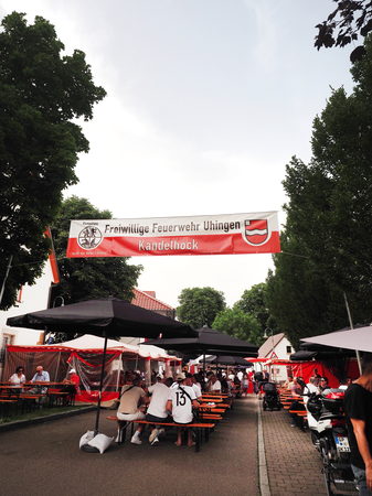 Unter einem zwischen Bäumen gespannten Banner mit der Aufschrift "Freiwillige Feuerwehr Uhingen Kandelhock" stehen Biertische und Bierbänke, auf denen Menschen sitzen.