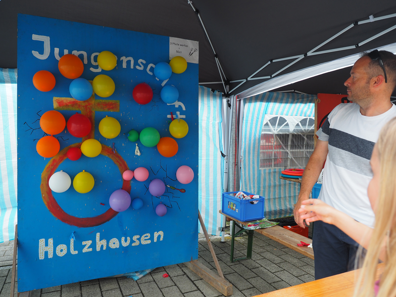 Unter einem Partyzelt stehe eine blaue Wand auf einem Biertisch. An der Wand sind Luftballons befestigt, die mittels Dartpfeilen zum Platzen gebracht werden können.