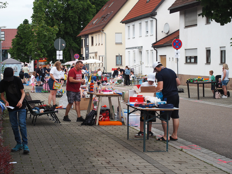 Am Rand einer Straße stehen Tische eines Kinderflohmarkts. Darauf sind Kinderspielzeuge und Kinderbücher. Menschen stehen vor den Ständen.