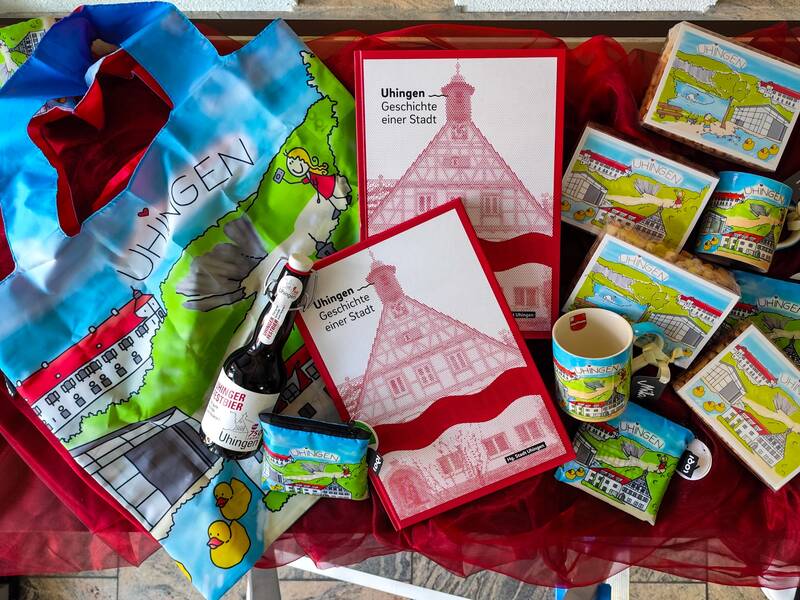 Auf einem roten Tuch sind die Uhinger Geschenkle abgebildet: Tasche, Bier, Buch, Wibele und Tasse