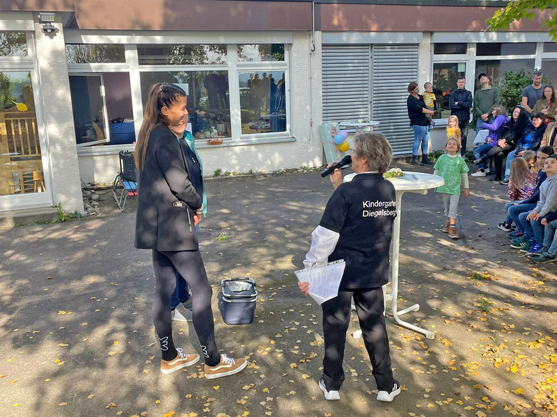 Eine Person hält vor einer kleinen Menschenmenge auf einem sonnigen Schulhof eine Rede.