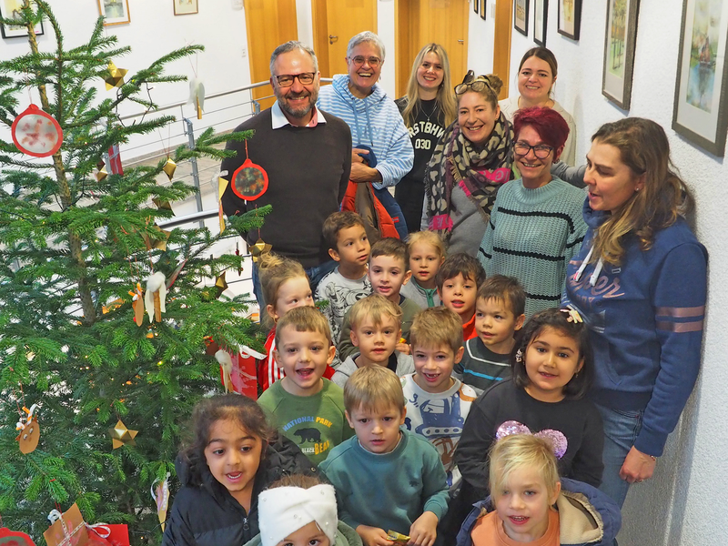 Eine Gruppe von Kindern und Erwachsenen steht neben einem geschmückten Weihnachtsbaum in einem Flur. Nicht nur Bürgermeister Matthias Wittlinger (hinten links) und das Kindergartenteam um Abteilungsleiterin Anette Epping (hinten, Zweite von links) mit (hinten, von links) Celina Haselwander und Yvonne Klockow erfreuen sich am Anblick des Baumes, auch Besucherinnen sowie Besucher.