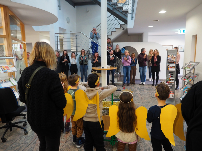 In einem Foyer stehen mehrere Kinder mit gelben Flügeln vor einer Gruppe applaudierender Erwachsener.