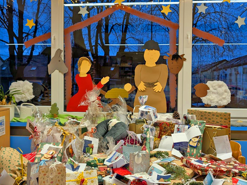 Ein Tisch voller bunter Geschenke steht vor einem Fenster, das mit einer Weihnachtskrippe aus Papier und Lichterketten dekoriert ist.