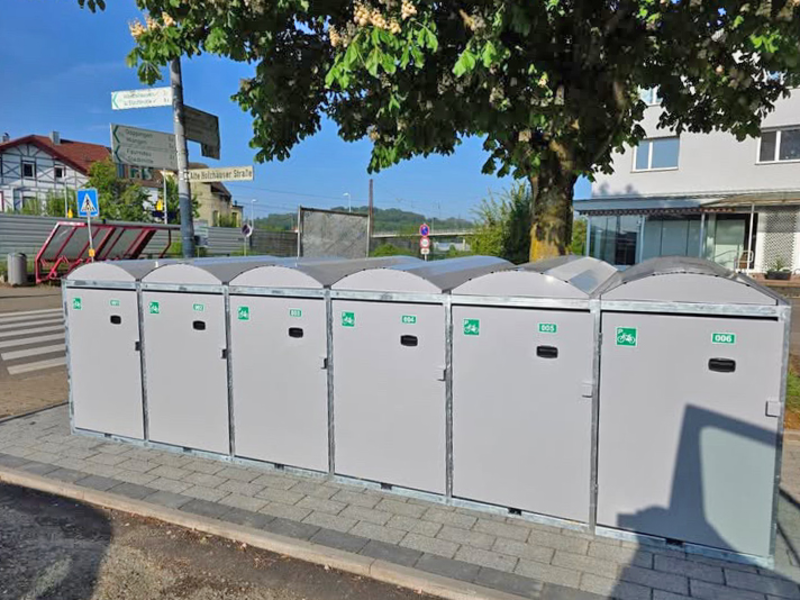 Eine Reihe grauer Container für Fahrräder steht ordentlich auf einem gepflasterten Gehweg unter einem Baum neben der Eisenbahnstraße beim Uhinger Bahnhof.