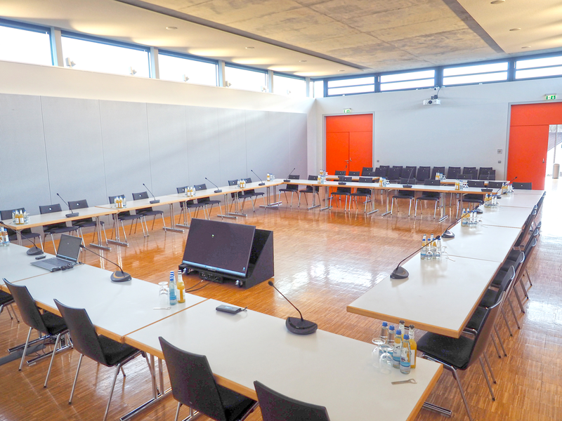 Ein heller Konferenzraum (der kleine Sitzungsaal im Uditorium) mit langen, zu einem U geformten Tischen, auf denen Mikrofone, Flaschen und Gläser stehen, und im Hintergrund befinden sich rote Türen.
