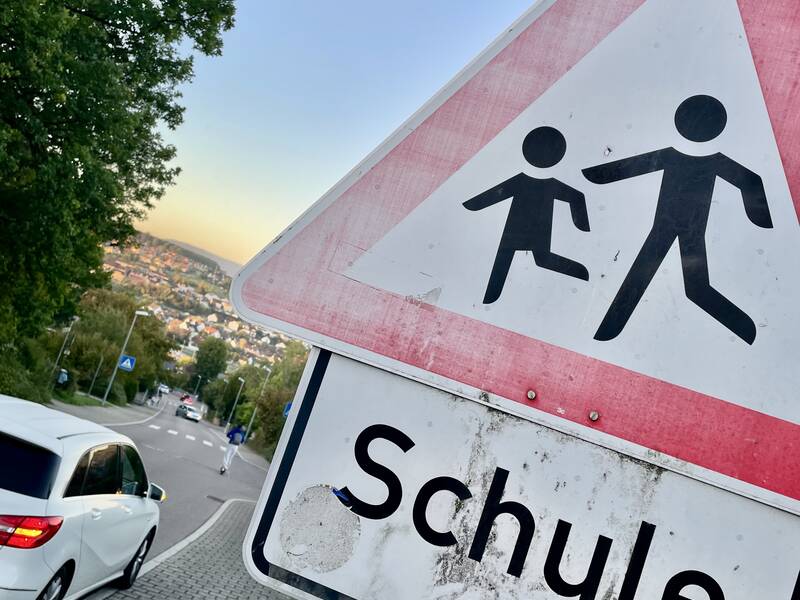 Ein Verkehrsschild mit der Aufschrift "Schule" zeigt eine Warnung vor Kindern, die eine Straße überqueren, während ein Auto auf einer kurvigen Straße fährt.