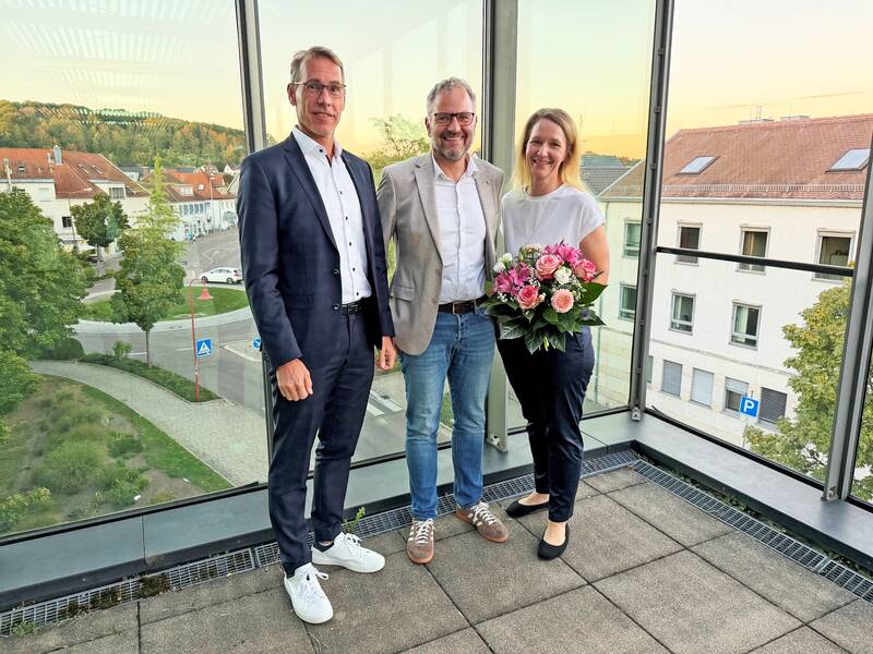 Drei Personen stehen auf einer Terrasse mit einer Glasbrüstung, wobei die Frau rechts einen Blumenstrauß hält und im Hintergrund eine Stadtlandschaft zu sehen ist.