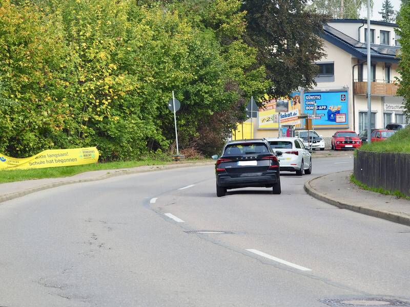 Mehrere Autos fahren auf einer Straße entlang, die von Bäumen und einem Haus mit Werbetafeln gesäumt ist, neben einem gelben Banner, das Autofahrer zur Vorsicht ermahnt, da die Schule begonnen hat.