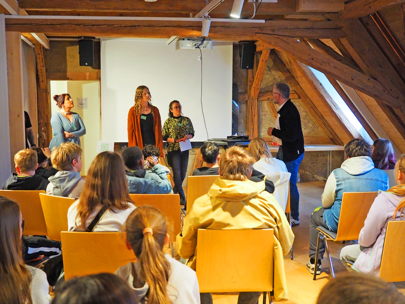 Eine Gruppe von Menschen sitzt in einem Raum mit Holzbalken, während vier Personen vorn stehen und scheinbar eine Präsentation halten.