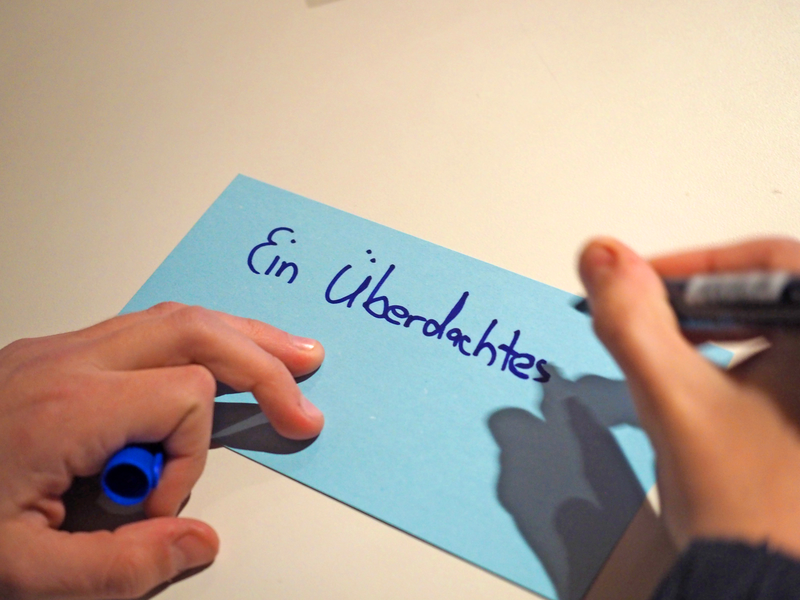 Zwei Hände halten und schreiben mit einem blauen Stift auf einer blauen Karteikarte den Text "Ein Überdachtes".