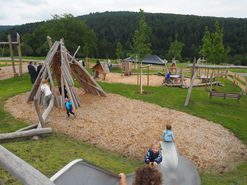 Der neue Spielplatz in Diegelsberg besteht aus Holzstrukturen, auf dem Kinder spielen und Erwachsene am Rand stehen, ist in eine grüne Landschaft eingebettet.