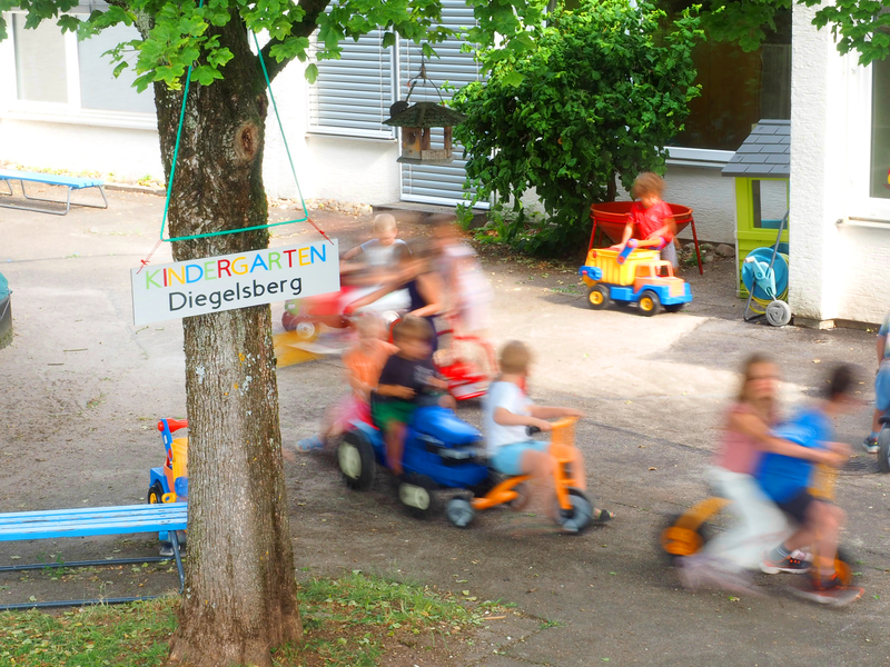 Mehrere Kinder fahren im Kindergarten Diegelsberg auf verschiedenen bunten Spielzeugfahrzeugen über einen gepflasterten Platz neben einem Schild mit der Aufschrift "KINDERGARTEN Diegelsberg".